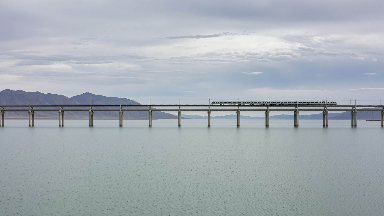 A long low rail causeway running straight out across flat grey open water under an overcast sky, distant mountains beyond.